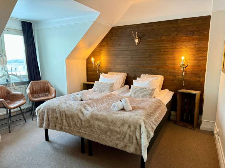Das Bild zeigt ein Schlafzimmer mit einem Doppelbett, zwei Sesseln und einem Fenster mit Vorhängen.