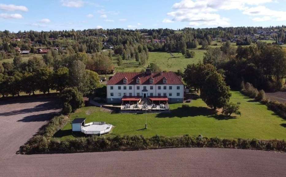 Ein großes, weißes Gebäude mit roten Dächern steht auf einem grünen Rasen vor einer bewaldeten Hügellandschaft.