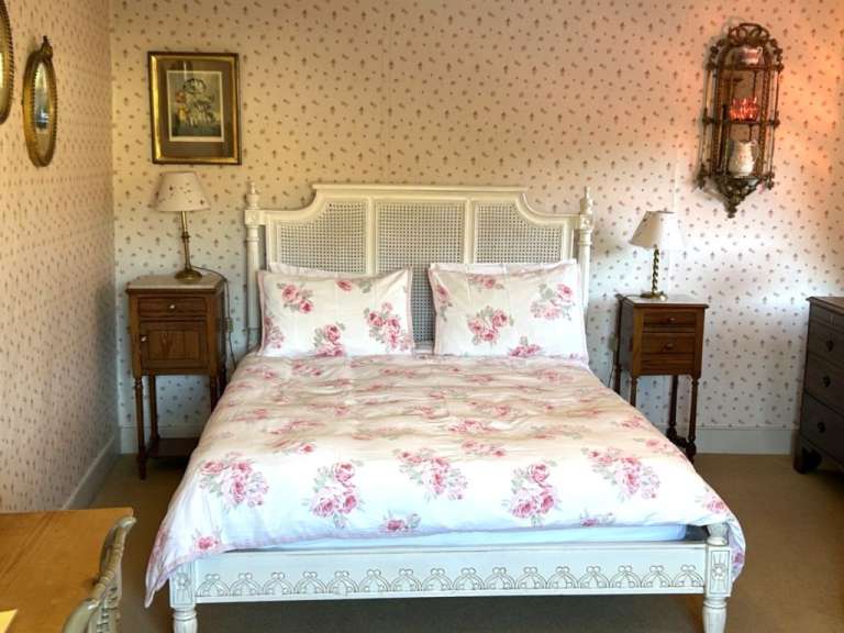 Ein Doppelbett mit floraler Bettwäsche steht in einem Raum mit blumiger Tapete, Nachttischen, Lampen und einem Wandregal.