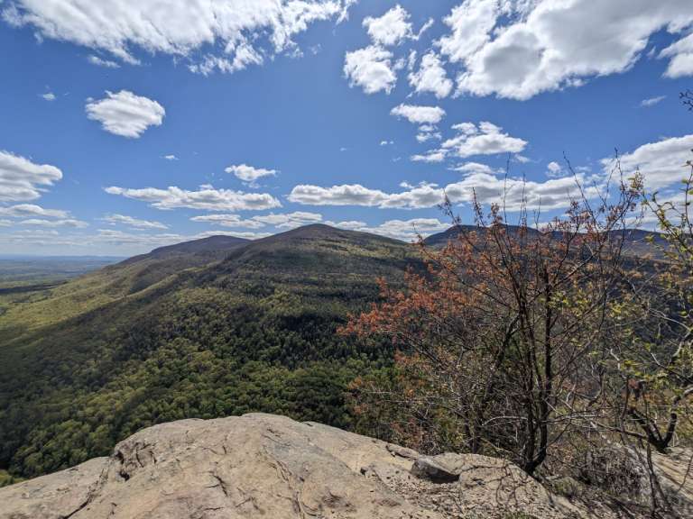 Eine weite Berglandschaft mit bewaldeten Hängen und felsigen Vordergrund unter einem blauen Himmel mit Wolken.