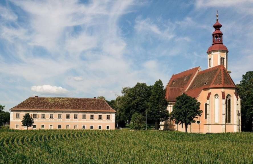 Ein zweistöckiges Gebäude und eine Kirche mit rotem Dach und Turm stehen vor einem Feld mit jungem Getreide unter einem blauen Himmel mit Wolken.