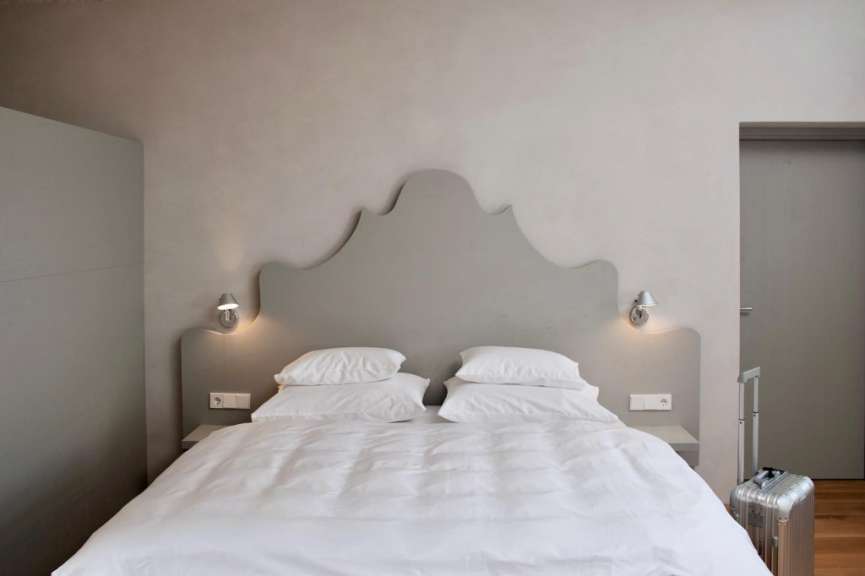 Ein Hotelbett mit weißen Kissen und Decken steht vor einem auffälligen Kopfteil an einer grauen Wand, daneben ein Koffer.