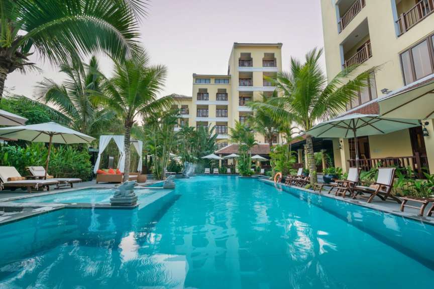 Ein tropischer Hotelpool ist von Palmen und Liegestühlen umgeben, während im Hintergrund ein mehrstöckiges Gebäude zu sehen ist.