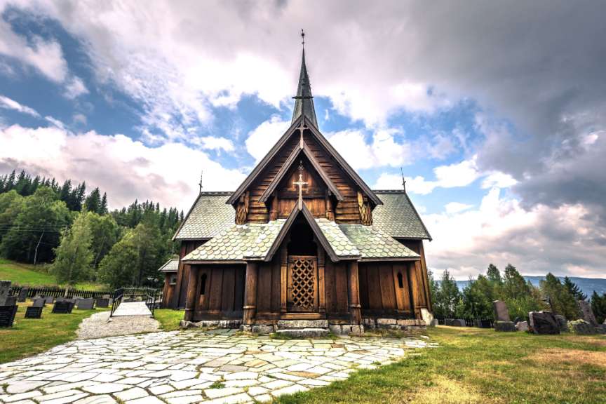 Eine Holzstabkirche mit einem spitz zulaufenden Kirchturm und geschwungenen Dächern steht vor einem bewaldeten Hügel und einem dramatischen Himmel.