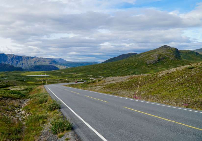 Ein kurvenreicher asphaltierter Weg schlängelt sich durch eine grüne Hügellandschaft mit zerklüfteten Bergen im Hintergrund unter einem wolkenverhangenen Himmel.