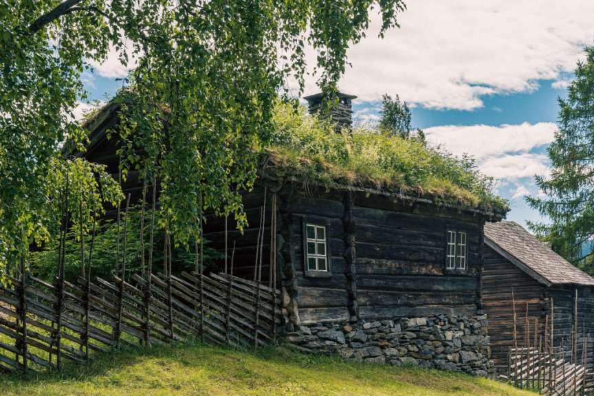 Ein altes Holzhaus mit einem begrünten Dach und einem Steinzaun steht auf einem Hügel unter einem blauen Himmel mit Wolken.