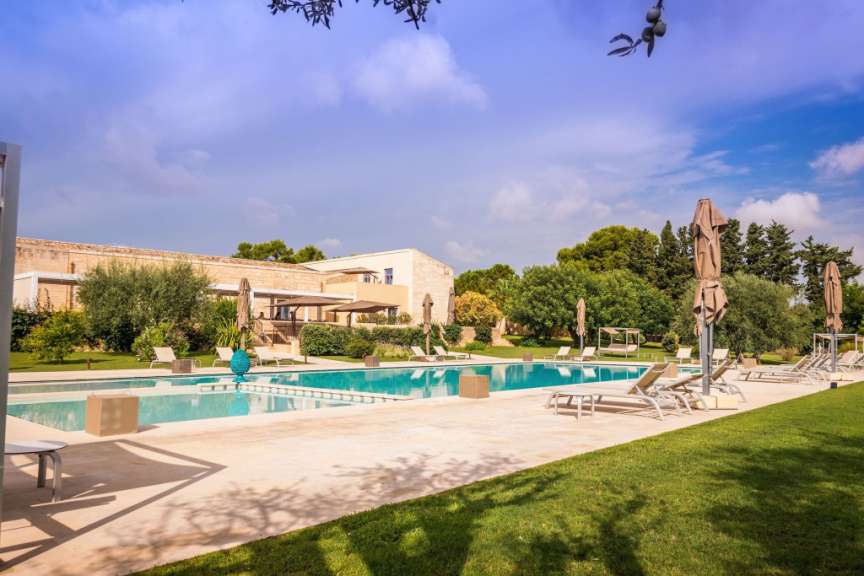 Ein großer Swimmingpool mit Liegestühlen und Sonnenschirmen steht im Schatten von Bäumen und einem Gebäude.