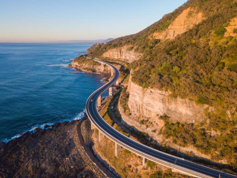 Ein Flugzeugflug über eine Kurve auf einer Autobahn mit Blick auf das Meer und einen Berg.