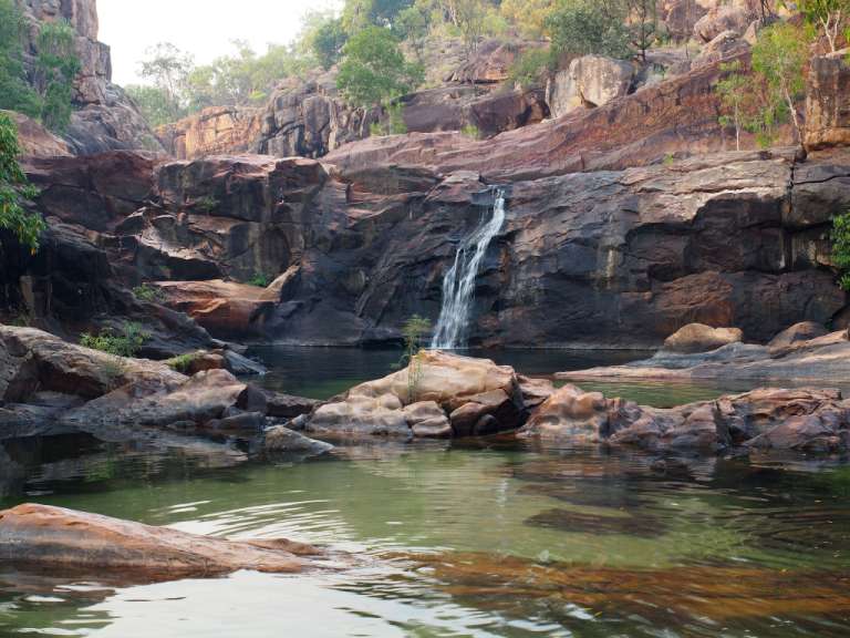 Ein schmaler Wasserfall stürzt von einer dunklen Felswand in ein Becken mit grünem Wasser.