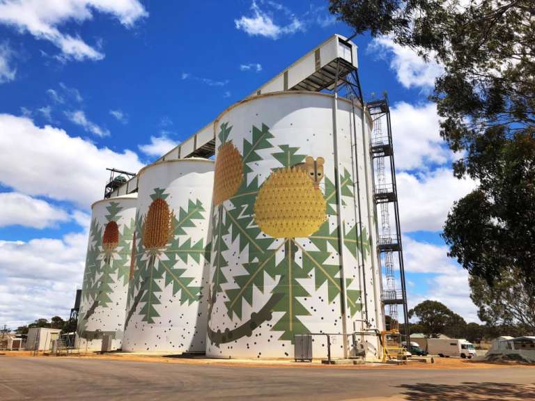 Große, bemalte Silos im Freien mit einem Banded-Banksia-Baummotiv gegen einen blauen Himmel mit Wolken.
