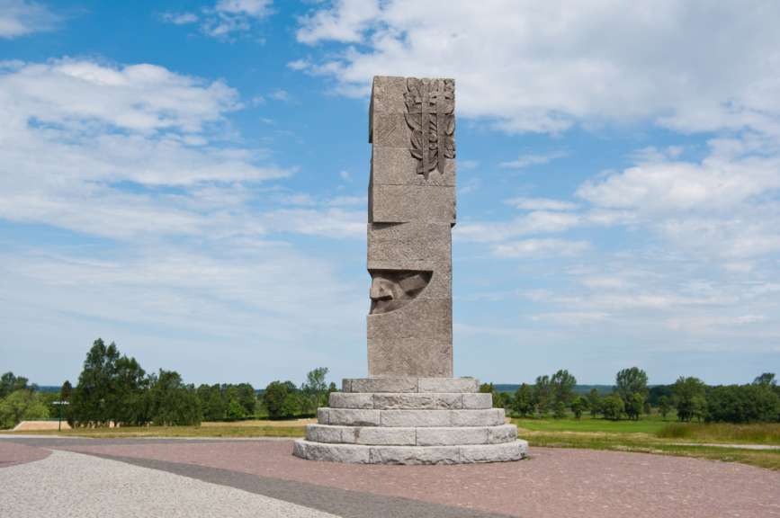 Eine stilisierte Steinstatue mit mehreren Stufen davor steht vor einem blauen Himmel mit Wolken und Bäumen im Hintergrund.
