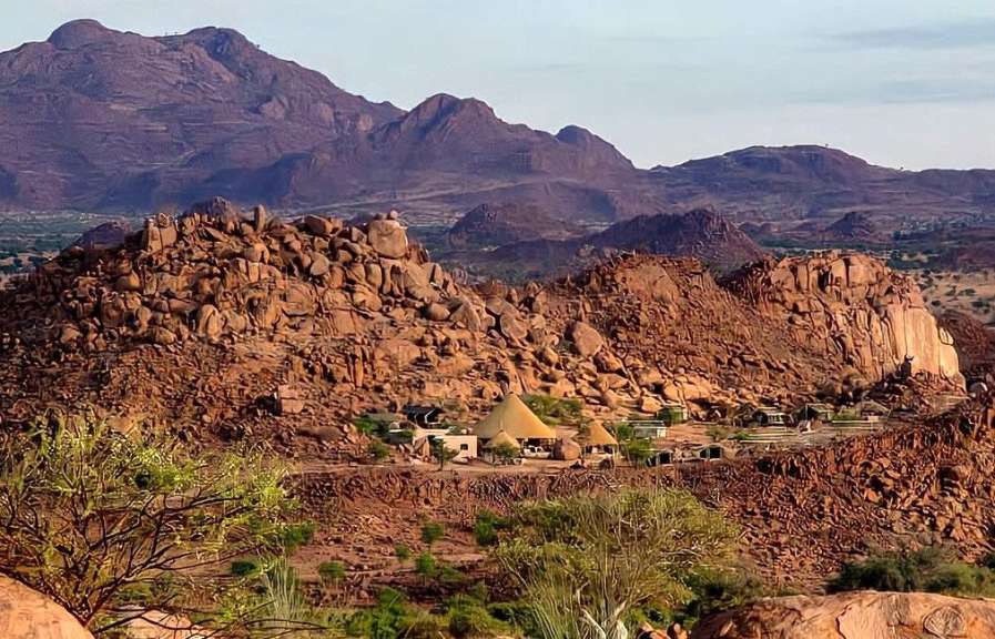 Ein Dorf mit traditionellen Behausungen und Zelten liegt inmitten einer felsigen und bergigen Landschaft mit vereinzelten Bäumen.