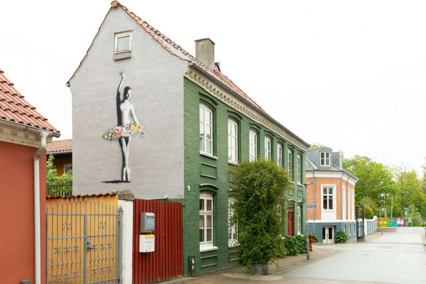 Ein Haus mit einem Graffito einer Ballerina steht auf einer Straße mit anderen Gebäuden und Bäumen.