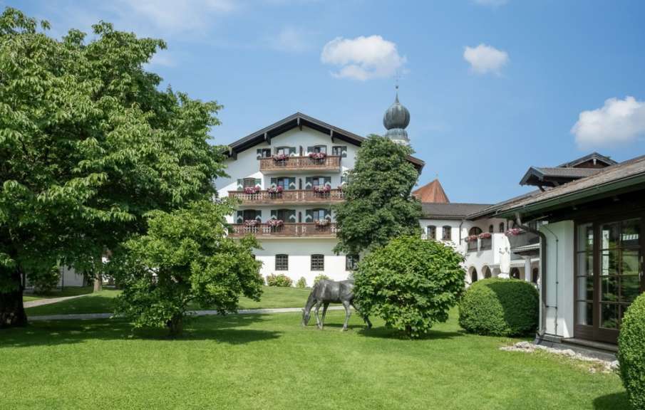 Ein Gebäude im alpinen Stil steht auf einer grünen Rasenfläche mit Bäumen, einem Pferde-Skulptur und einem bewölkten Himmel.