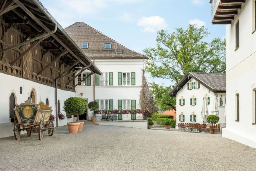 Ein Innenhof mit einem alten Pferdekarren, einem großen Haus mit grünen Fensterläden, einem kleineren Gebäude mit einer Außenterrasse und einem großen Baum.