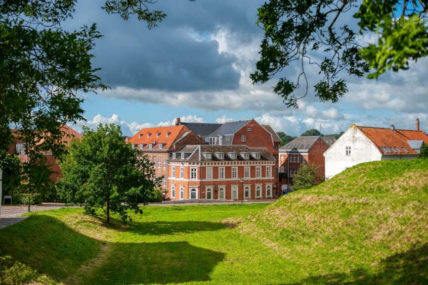 Grüne Hügel führen den Blick zu roten und weißen Gebäuden, die von Bäumen und einer dramatischen Wolkendecke eingerahmt sind.