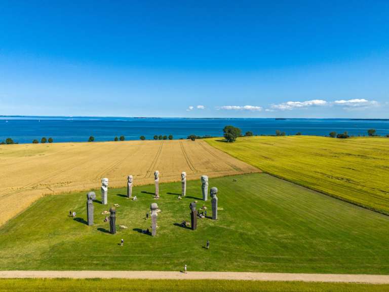Ein Blick von oben auf eine Landschaft mit einem Feld, einer Rasenfläche mit Steinskulpturen und dem Meer im Hintergrund.