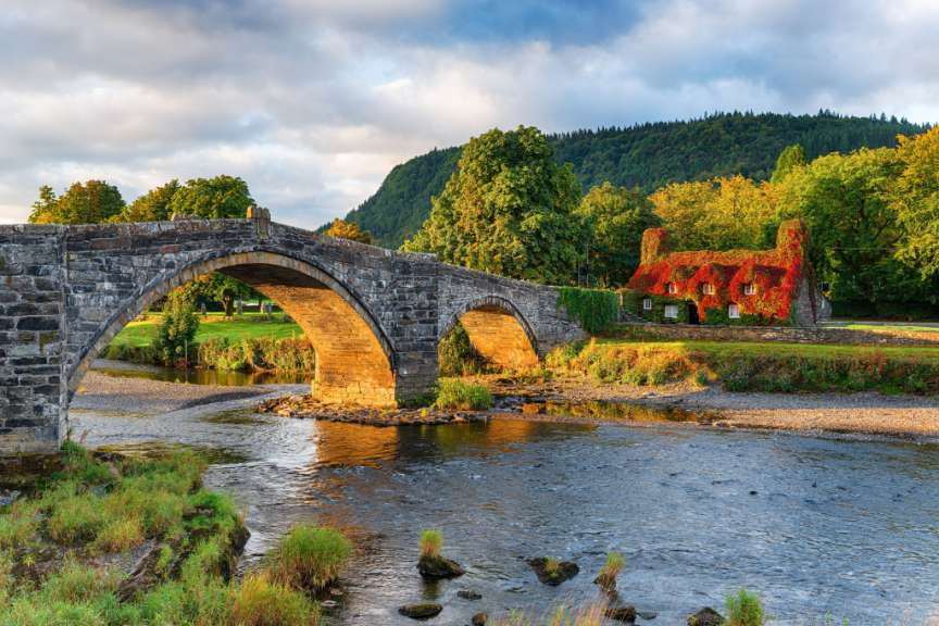 Eine alte Steinbrücke überspannt einen Fluss vor einem bewaldeten Hügel, neben einem Haus, das mit rotem und grünem Laub bedeckt ist.