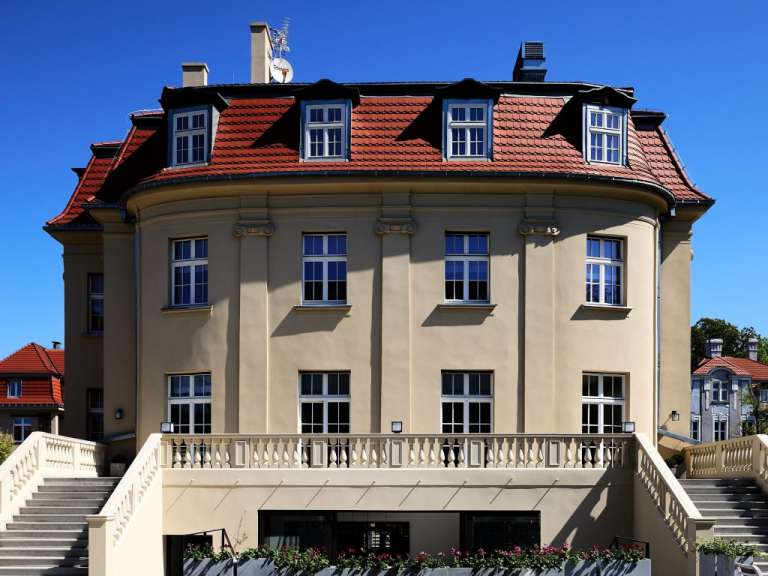 Eine Ansicht eines großen, hellgelben Gebäudes mit rotem Ziegeldach und einer breiten Steintreppe, die zu einer Balustrade führt, vor einem strahlend blauen Himmel.