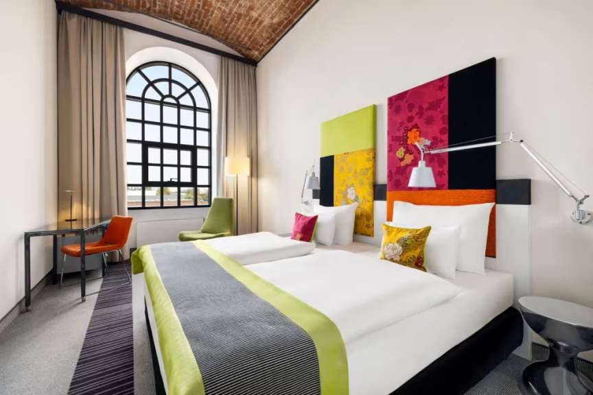 Das Bild zeigt ein stilvolles Hotelzimmer mit zwei Betten, einem großen Bogenfenster und einem architektonischen Backsteingewölbe.