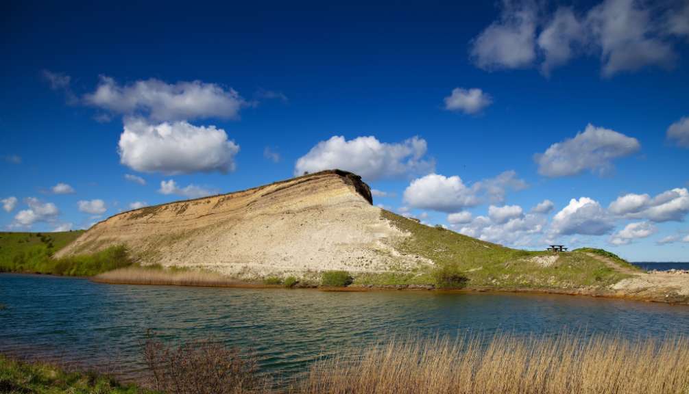 Ein großer, sandiger Hügel mit einer Klippe ragt über einem ruhigen See auf, unter einem dramatischen blauen Himmel mit dramatischen Wolken.
