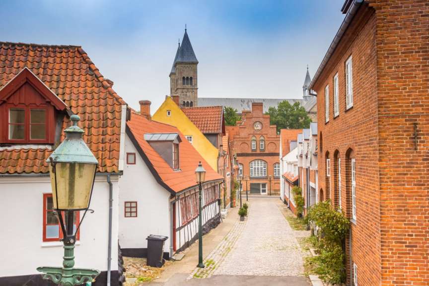 In einer europäischen Stadt führt eine Kopfsteinpflasterstraße zwischen bunten Häusern hindurch, während im Hintergrund eine Kirche mit zwei Türmen aufragt.
