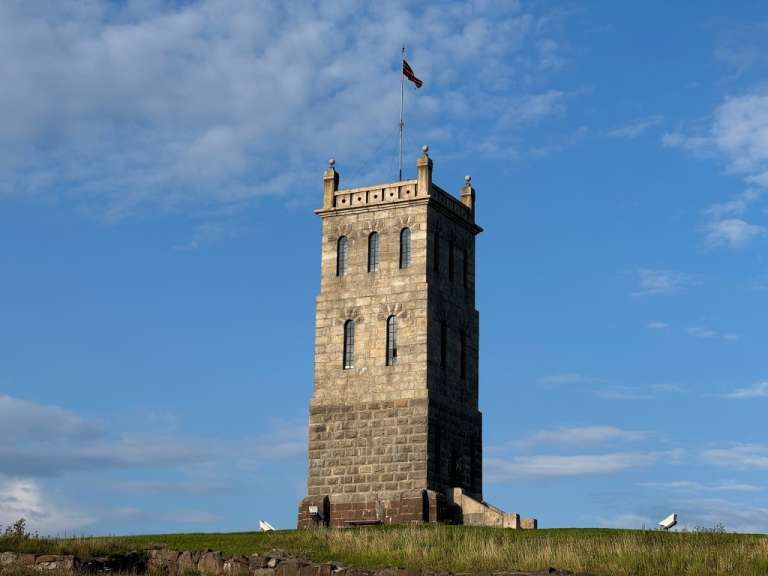 Eine Steinturm mit einem norwegischen Flagge im oberen Teil und auf einem Grasberg unter blauem Himmel mit Wolken.