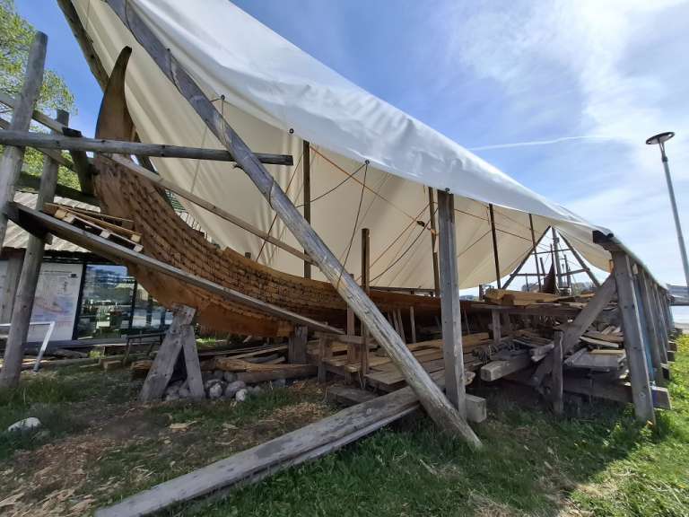 Ein Holzschiff wird unter einem großen weißen Tarp auf einer Holzkonstruktion restauriert.