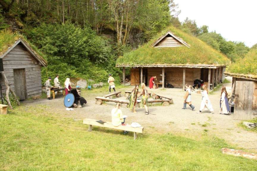 Mehrere Menschen in historischen Kostümen sind in einer Freilichtanlage mit Holzgebäuden und Grasdächern zu sehen.