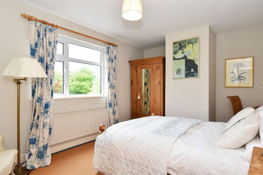 Ein gemütliches Schlafzimmer mit einem Doppelbett, einem Holzkleiderschrank, einem Fenster mit floralen Vorhängen und zwei gerahmten Bildern an der Wand.