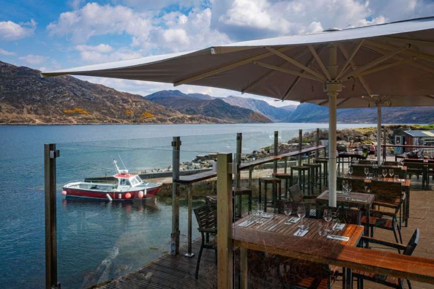 Eine Terrasse mit Tischen und Stühlen blickt auf einen Fjord mit einem Boot, während im Hintergrund Berge aufragen.