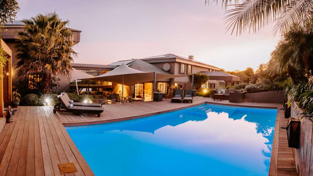 Ein luxuriöser Außenbereich mit einem Pool, Liegestühlen und einer überdachten Terrasse wird bei Sonnenuntergang von warmem Licht beleuchtet.