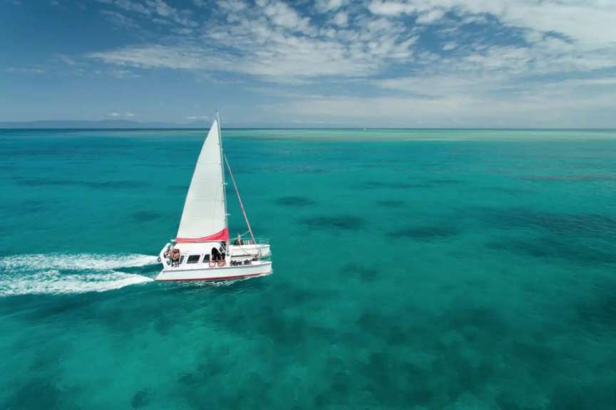 Ein Segelkatamaran fährt durch türkisfarbenes Wasser unter einem bewölkten blauen Himmel.