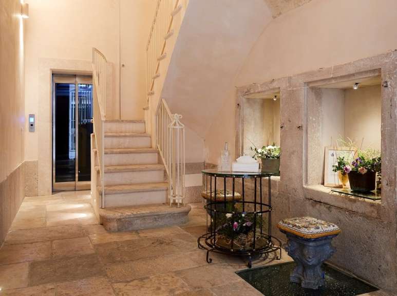 Ein heller Flur mit Steinböden, einer Treppe, einem Aufzug und dekorativen Nischen mit Blumenarrangements.