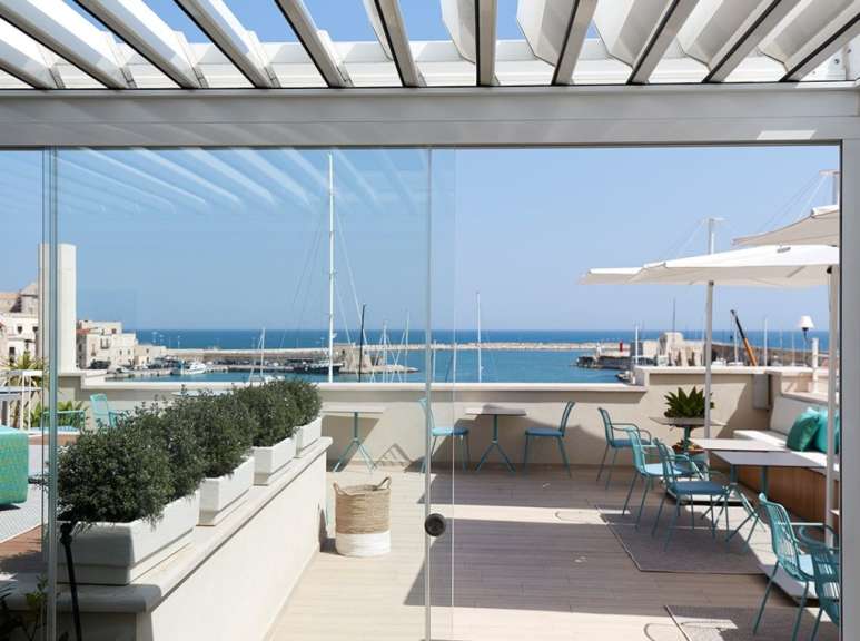 Auf einer Terrasse mit Blick auf das Meer und einen Hafen stehen blaugrüne Stühle und Tische unter weißen Sonnenschirmen.
