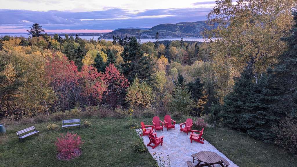 Ein Blick von einem erhöhten Aussichtspunkt auf eine von bunten Herbstbäumen gesäumte Landschaft mit einem Gewässer und Hügeln im Hintergrund, wobei im Vordergrund ein Sitzbereich mit roten Adirondack-Stühlen und einer Parkbank zu sehen ist.