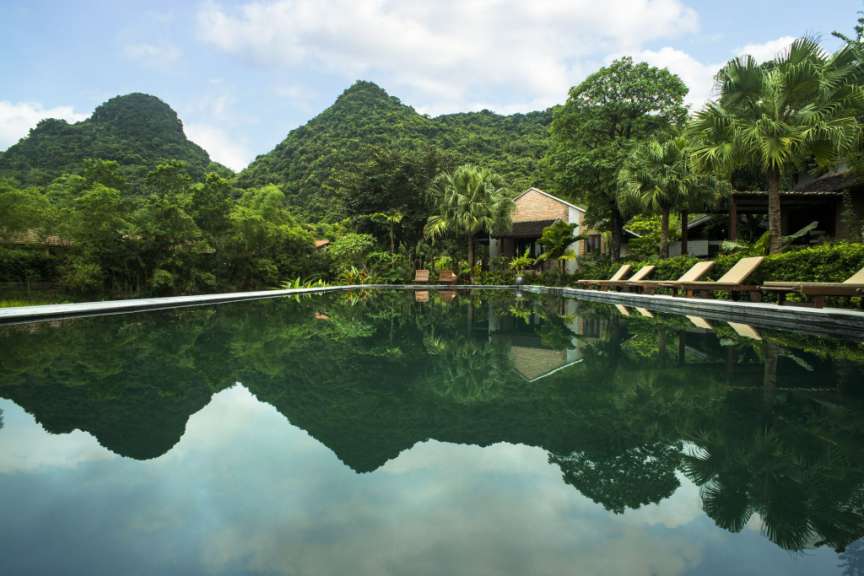 Ein Infinity-Pool spiegelt bewaldete Berge und ein Gebäude in einer tropischen Landschaft.