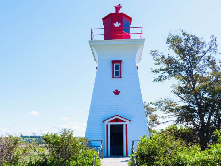 Ein weißer Leuchtturm mit rotem Dach und einer kanadischen Flagge steht an einem sonnigen Tag auf einem Hügel, umgeben von grünen Bäumen und Büschen.