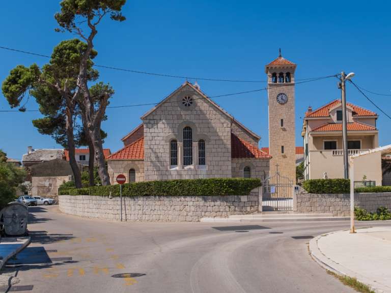 Ein zweistöckiges Gebäude mit orangefarbenem Dach steht neben einem Kirchturm mit Uhr auf der linken Seite eines breiten, asphaltierten Weges.