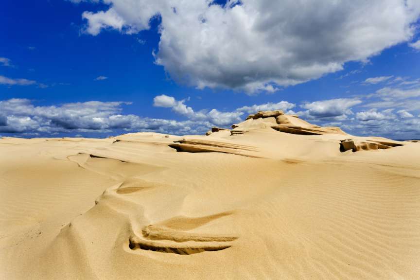 Das Bild zeigt eine weite Sandlandschaft mit welligen Dünen unter einem strahlend blauen Himmel mit weißen Wolken.