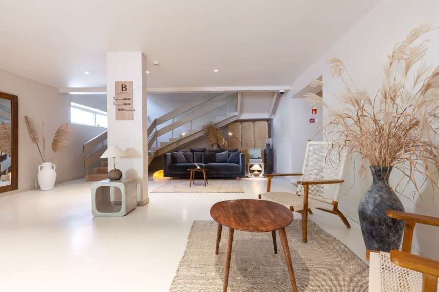 Eine helle Lobby mit einem dunklen Sofa, zwei Holzstühlen, einem Couchtisch, einem großen Krug mit getrockneten Gräsern und einer Treppe im Hintergrund.