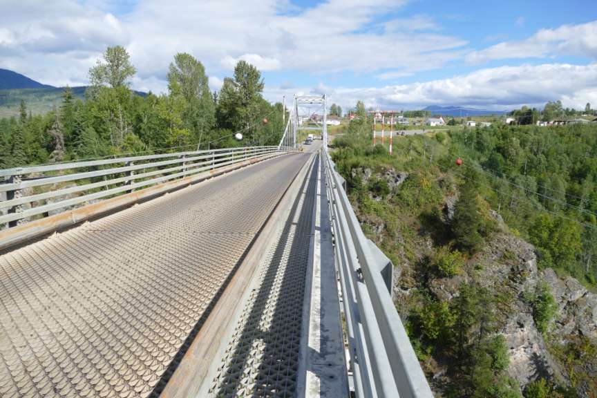 Eine Brücke aus Metallgitter überquert eine felsige Schlucht, auf der sich Bäume und Häuser mit roten Dächern und weißen Wänden befinden.