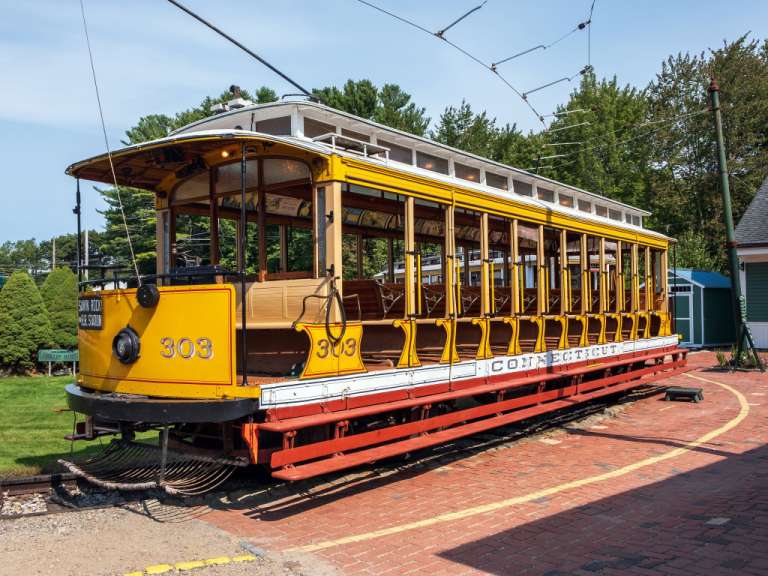 Ein gelber Oldtimer-Trolley mit der Nummer 303 steht auf einem roten Backsteinbahnsteig vor Bäumen und blauer Himmel.