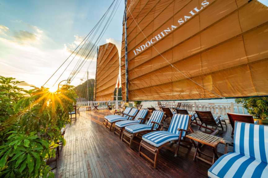 Liegestühle und Tische auf einem Holzdeck neben einem Segelboot mit der Aufschrift indochina sails