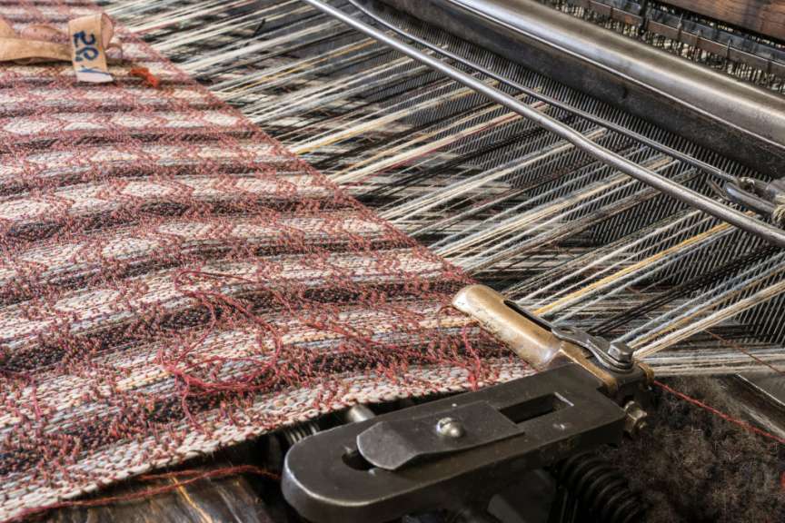 Eine Nahaufnahme eines Webstuhls, der ein rot-weißes gemustertes Textil mit einer Vielzahl von bunten Fäden in verschiedenen Stadien der Weberei zeigt.