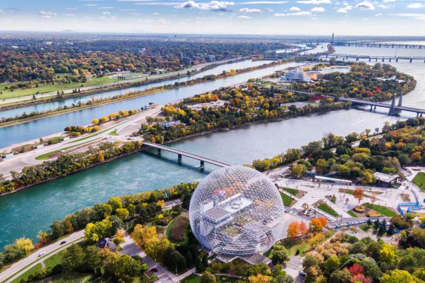 Ein breiter Fluss mit einer Stadtlandschaft und vielen Brücken im Hintergrund, während im Vordergrund ein großes, kuppelförmiges Gebäude aus Gitterwerk zu sehen ist.