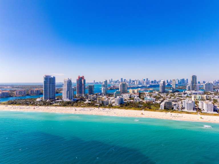 Eine Drohnenaufnahme zeigt eine belebte Stadtlandschaft mit hoch aufragenden Gebäuden, einem goldenen Strand mit vielen Menschen und dem türkisblauen Wasser des Ozeans.