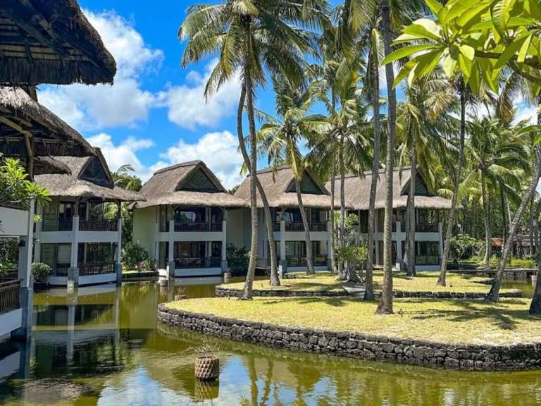 Ein tropischer Resort mit strohgedeckten Gebäuden und Kokospalmen, die sich in einem ruhigen Wasser spiegeln.