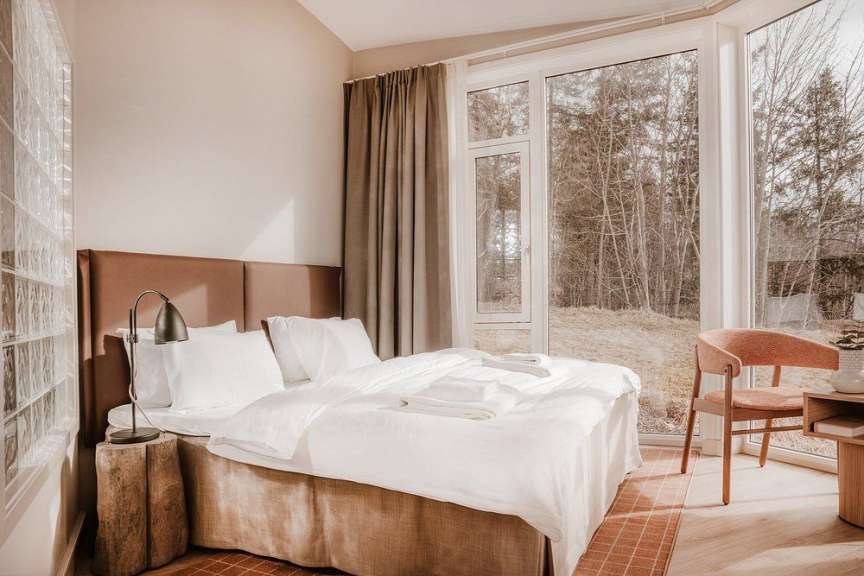 Das Bild zeigt ein Bett in einem Raum mit großen Fenstern, die auf einen Wald blicken, daneben ein Stuhl und ein Nachttisch.