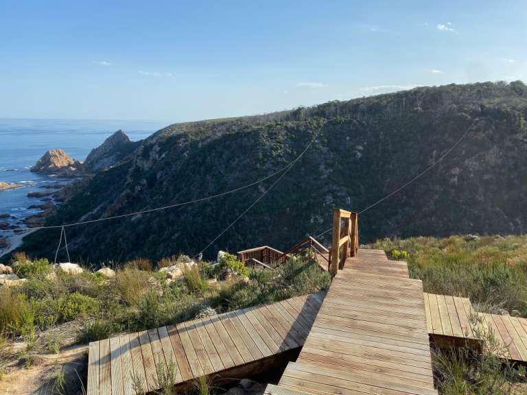 Ein Holzsteg führt hinunter zu einer Klippe mit Blick auf das Meer